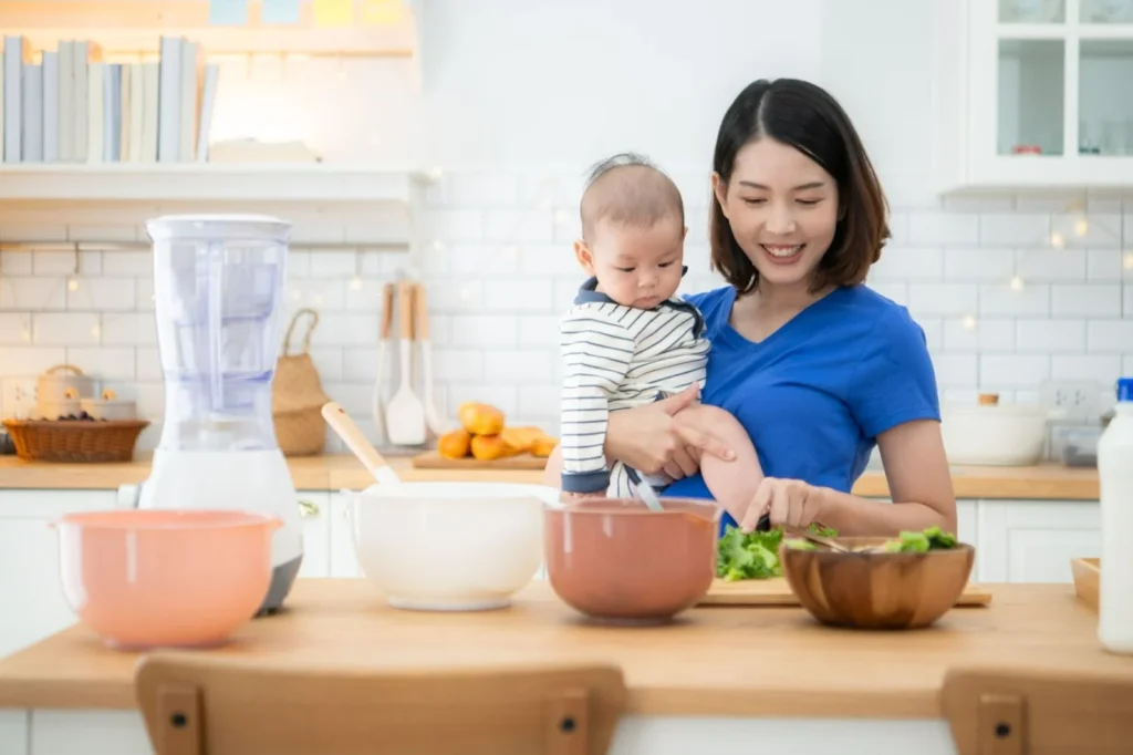 Postnatal mum preparing healthy food in the kitchen with her baby, representing the nutrition focus of confinement service Singapore care.