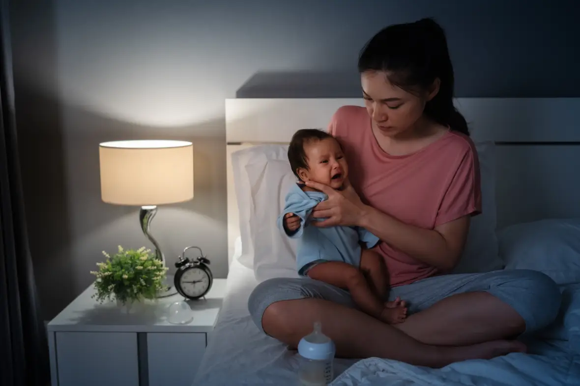 A mother settling in her baby for bed. The lamp is turned on and a baby bottle is in front of her, indicating that she's about to feed the baby.